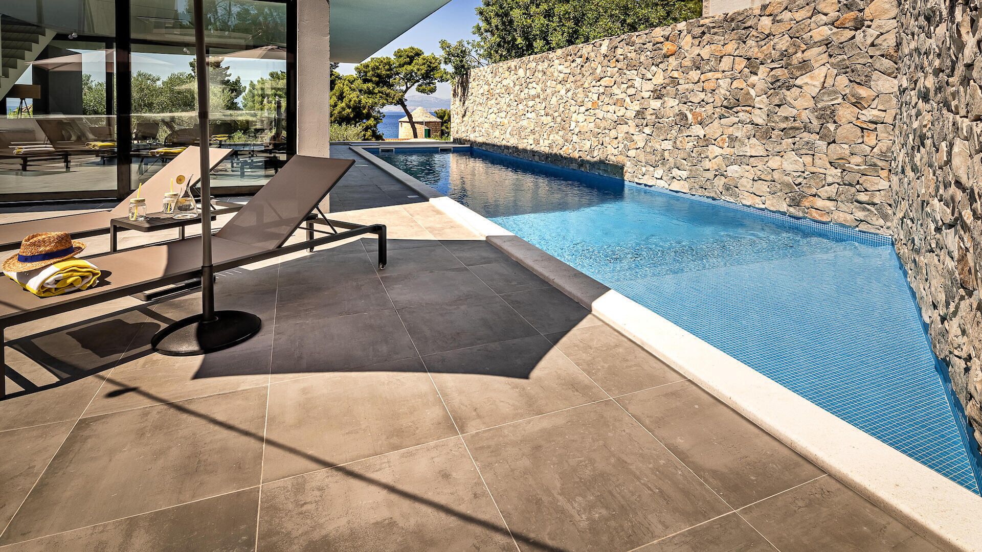 A modern outdoor pool area with stone walls, lounge chairs, and a glass-enclosed patio.