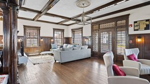 A spacious living room with a high ceiling, wooden beams, and large windows. There is a grey sofa, a white armchair, and a wooden cabinet.