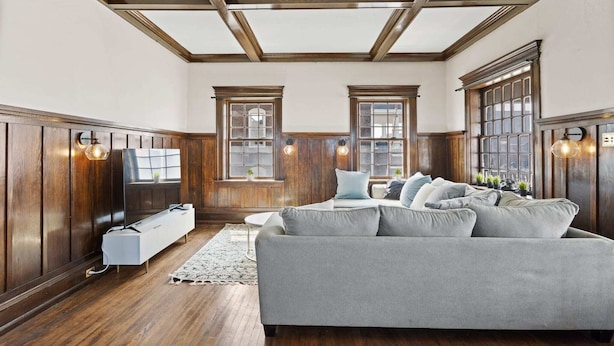 A living room with a grey sofa, wooden paneled walls, large windows, and a radiator.