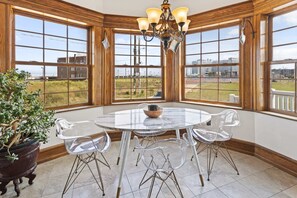 A dining area with a round table, clear chairs, a chandelier, and large windows offering a view of the outdoors.