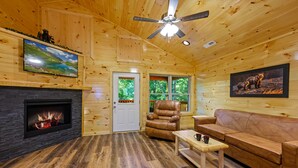 A log cabin interior with a fireplace, leather furniture, a ceiling fan, and framed artwork. - We love Dogs! Hot Tub, Pool Table, Media Room, Easy Access | Free Tickets! (Pigeon Forge)