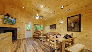 A wooden cabin interior with a fireplace, wooden furniture, a dining table, and a ceiling fan. - We love Dogs! Hot Tub, Pool Table, Media Room, Easy Access | Free Tickets! (Pigeon Forge)