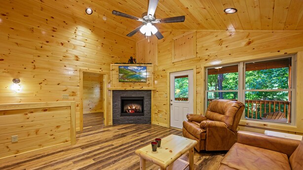A wooden cabin interior with a fireplace, leather seating, a television, and a balcony with a view of trees. - We love Dogs! Hot Tub, Pool Table, Media Room, Easy Access | Free Tickets! (Pigeon Forge)