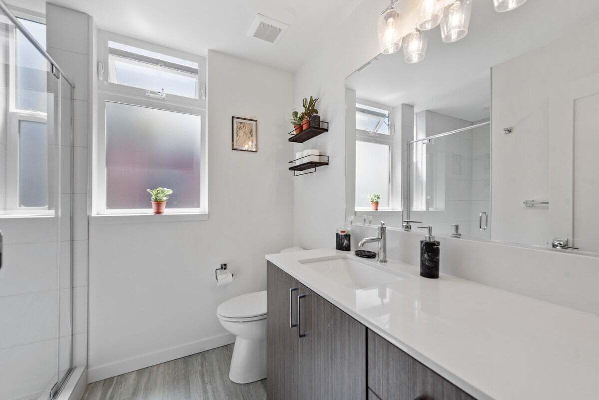 Un baño moderno con tocador blanco, un espejo grande y un inodoro. Dos ventanas dejan entrar luz natural, y hay una planta pequeña en el alféizar.