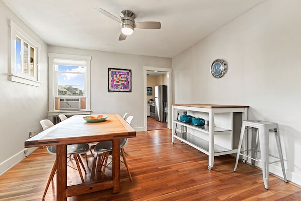 A dining area with a wooden table and chairs, a ceiling fan, and a view of the kitchen in the background.