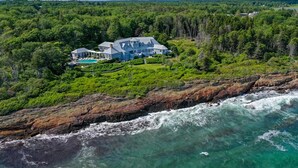 A large, two-story house with a swimming pool, surrounded by dense forest and overlooking a rocky coastline.