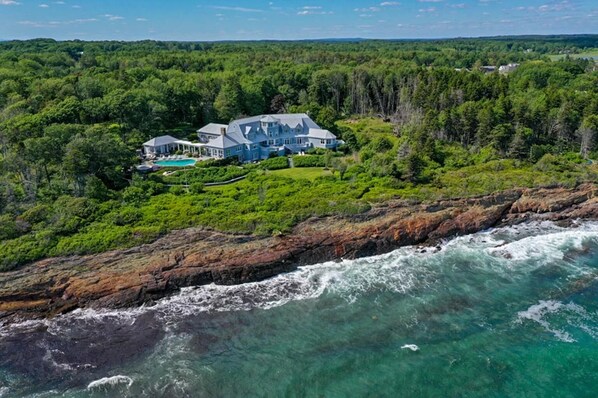 A large house with a pool, surrounded by dense forest and a rocky coastline.