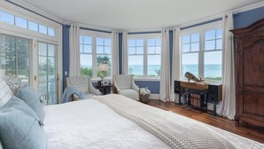 A bedroom with a large bed, two armchairs, a wooden dresser, and a view of the sea through large windows.