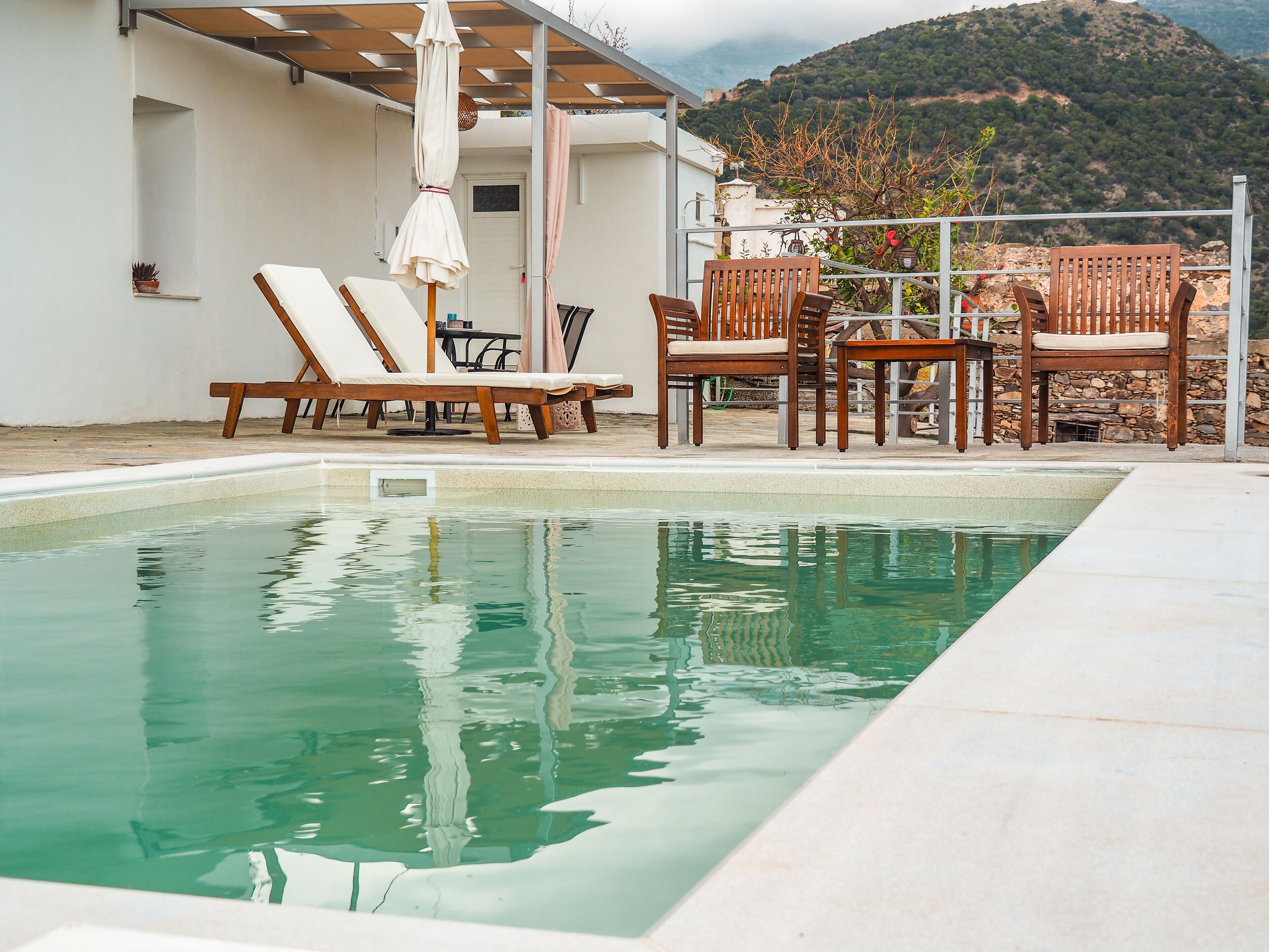 Ein Swimmingpool mit Liegestühlen und einem Tisch, umgeben von einem weißen Gebäude mit Blick auf eine gebirgige Landschaft.