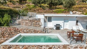 A pool area with stone walls, a white building with a door and windows, and outdoor furniture.