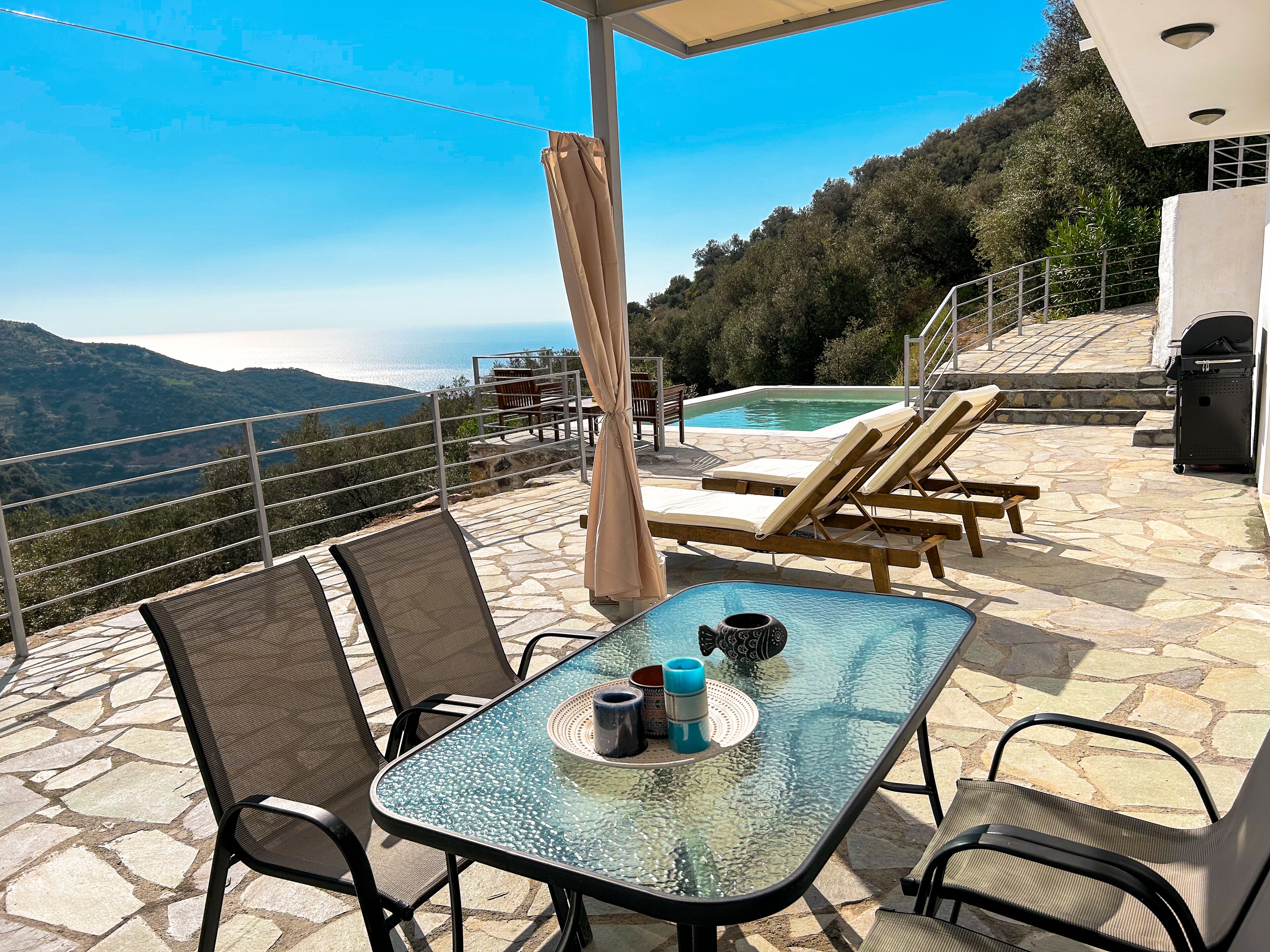 Een terras buiten met een glazen tafel, stoelen, een zwembad en uitzicht op de bergen.