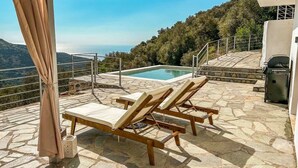 A poolside area with lounge chairs, a stone-tiled deck, and a view of the sea and hills.