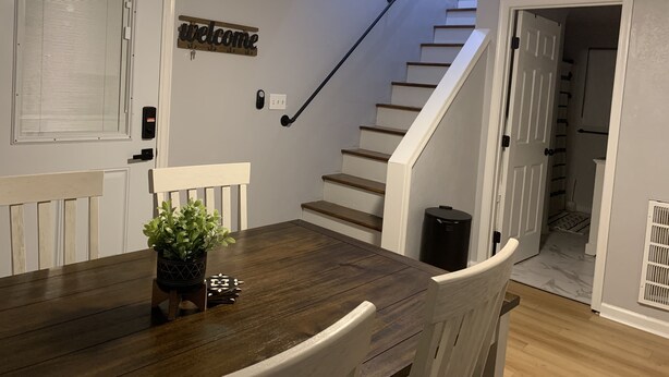 A dining area with a wooden table and chairs, a potted plant, a welcome sign, a staircase, and a door leading to another room.