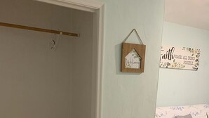 A room with a wooden floor, a bed with floral bedding, a wooden bench, a wall-mounted light fixture, and a small wooden sign with a house and the word "welcome".