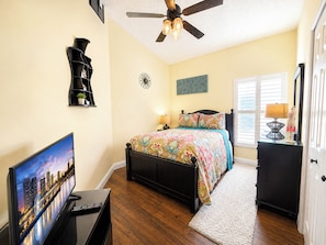 A bedroom with a bed, a TV, a ceiling fan, a window, and a door.