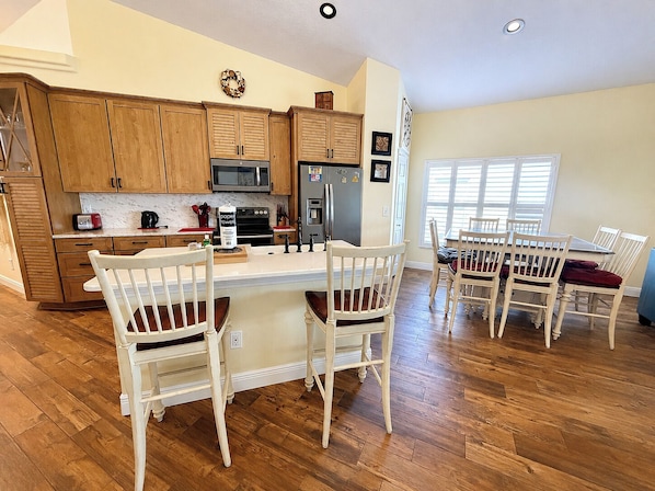 A kitchen with wooden cabinets, a central island with chairs, a dining table with chairs, and a refrigerator.