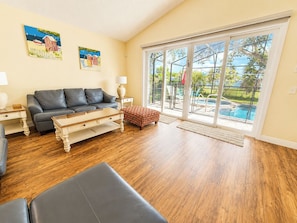A living room with a grey sofa, a wooden coffee table, and a patterned ottoman. There are two paintings on the wall, a sliding glass door leading to a pool area, and a wooden floor.