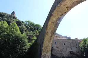 Un pont en pierre, dont l’arche s’étend au-dessus d’une colline verdoyante, avec un bâtiment historique en arrière-plan.