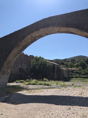 Un ancien pont de pierre en arc de cercle enjambant une rivière, avec un bâtiment rustique et de la verdure en arrière-plan.