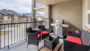 A balcony with wicker furniture, a grill, and a view of apartment buildings and mountains.