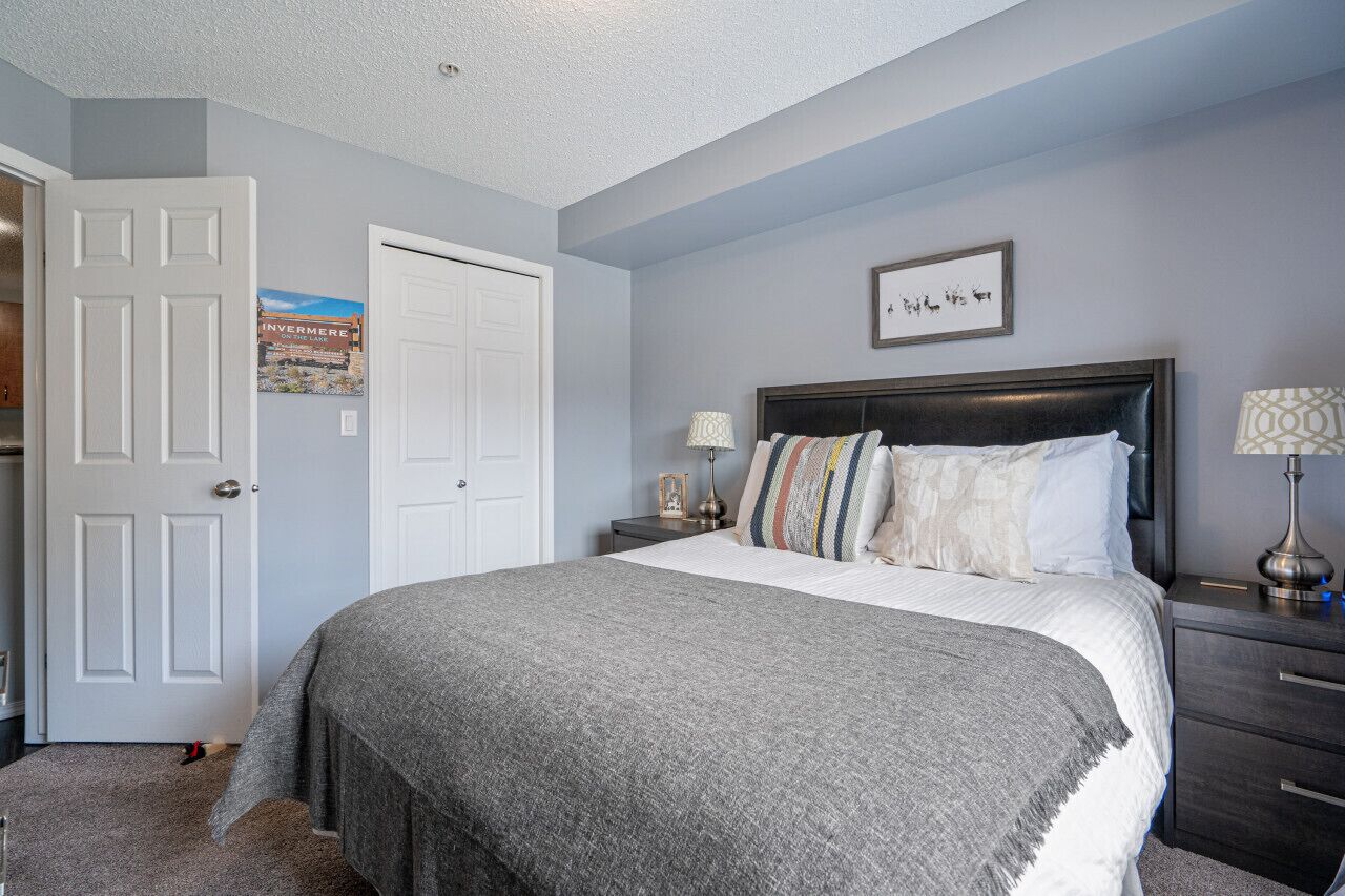 A bedroom with a large bed, a nightstand with a lamp, a framed picture on the wall, and a door with a poster.