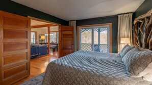 Ein Schlafzimmer mit einem großen Bett, Holzvertäfelung, Blick durch eine Schiebetür in einen Essbereich und einem großen Fenster mit Blick auf Bäume.