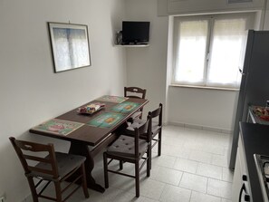 Un'area pranzo con un tavolo di legno, quattro sedie, un quadro appeso al muro, un televisore montato e una finestra con persiane.
