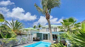 A pool area with a clear blue pool, surrounded by palm trees and a two-story building with a light blue roof. - Villa Zandoli – Private Pool Just Steps from Orient Bay Beach, 3 bedrooms (Orient Bay)