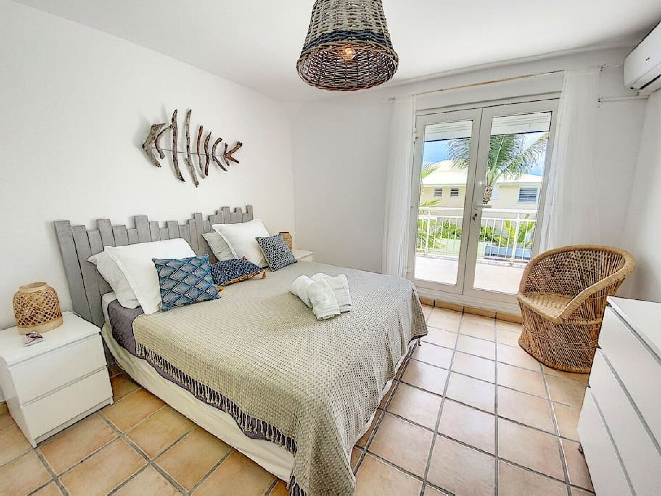 Une chambre à coucher comprenant un lit, des tables de chevet, un fauteuil en osier, un luminaire en fibres tressées et une vue sur l’extérieur.