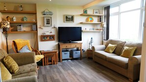 Ein Wohnzimmer mit einer Couch, Sesseln, einem Fernseher, Holzregalen und einem Fenster.