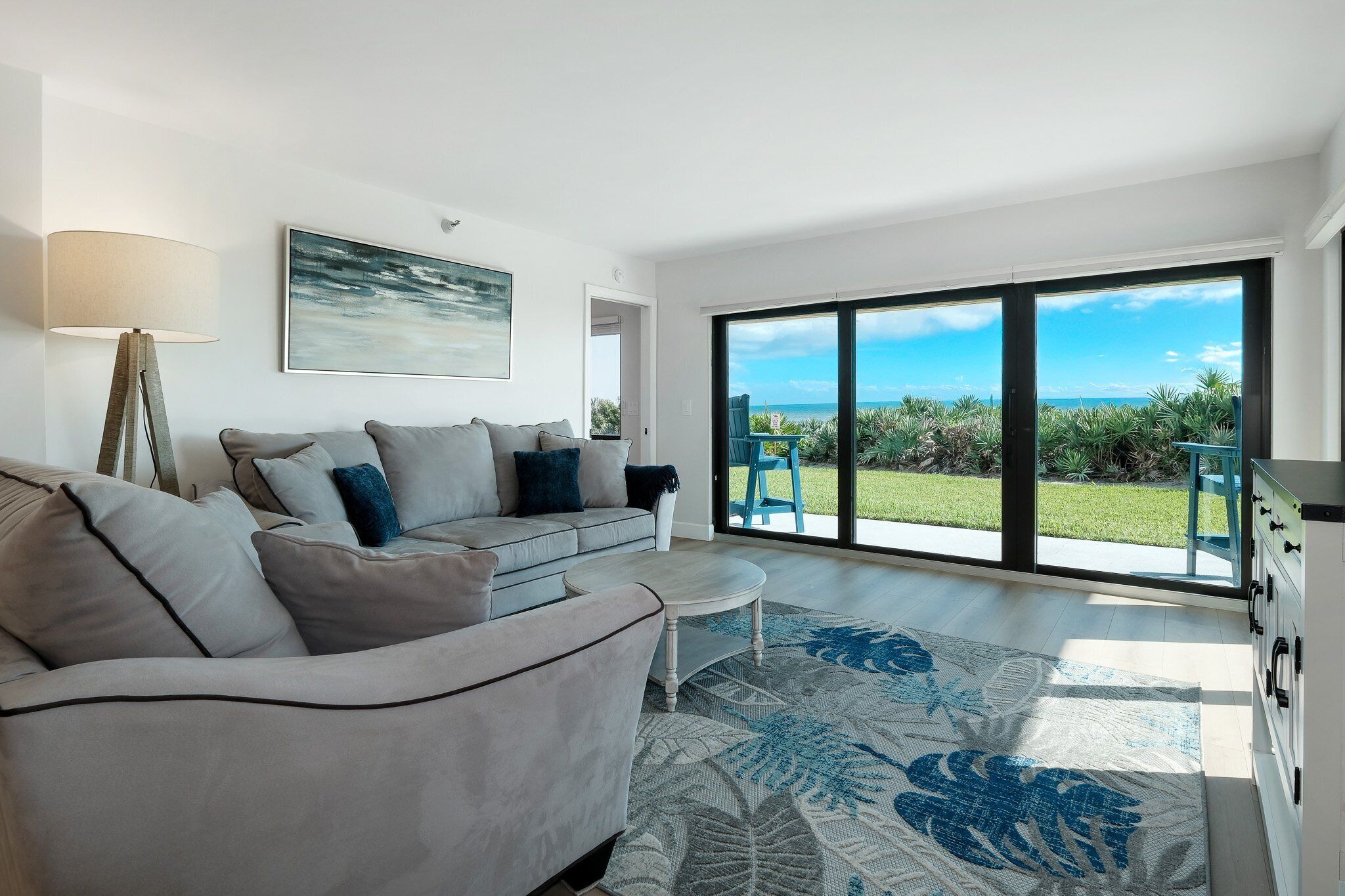 A modern living room with a grey sofa, a coffee table, and a large window offering a view of the sea and greenery.