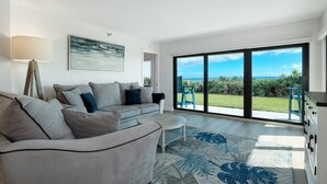 A modern living room with a grey sofa, a coffee table, and a large window offering a view of the sea and greenery.