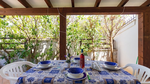 Un patio exterior con una mesa dispuesta para dos personas, cubierta por un mantel estampado, rodeado de sillas de plástico blanco.