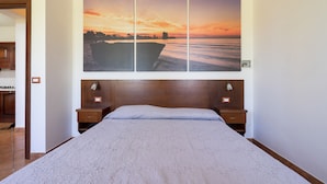 Un dormitorio con una cama grande, cabecera de madera, dos mesitas de noche y un cuadro colgado en la pared que muestra un atardecer sobre el agua.