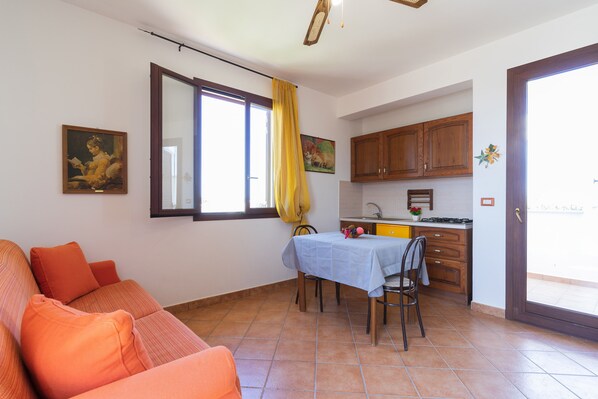 A living room with a sofa, a dining table with chairs, a kitchen area with cabinets, and a sliding glass door leading to an outdoor area.