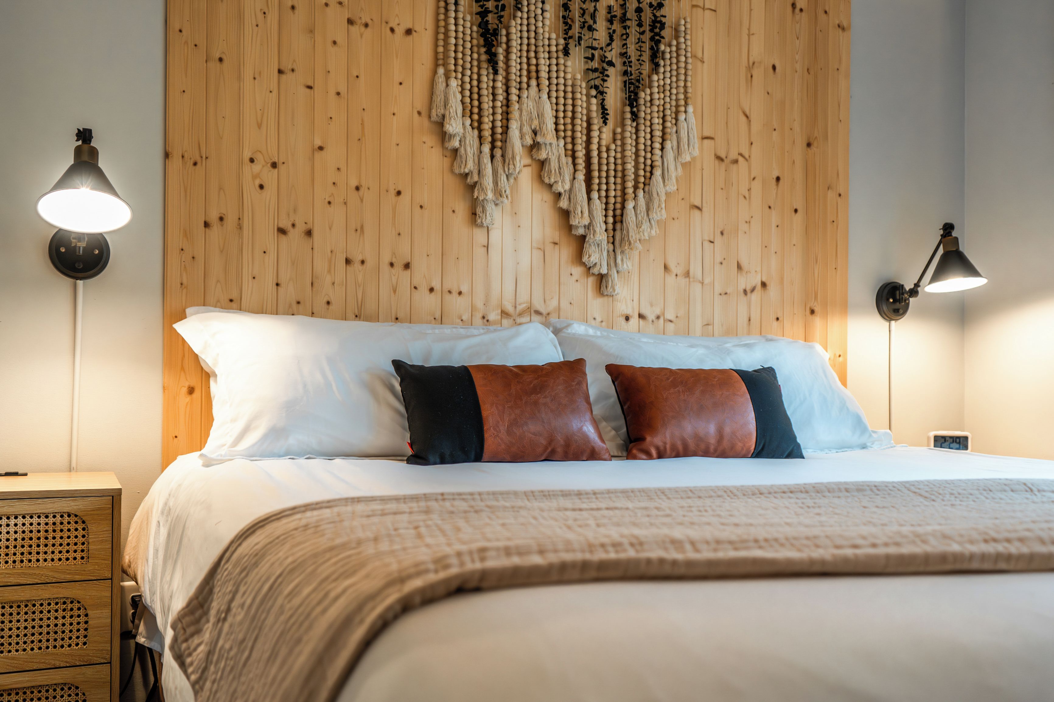 Una cama bien hecha con almohadas blancas y marrones, un cabecero de madera y un tapiz con borlas colgado en la pared.