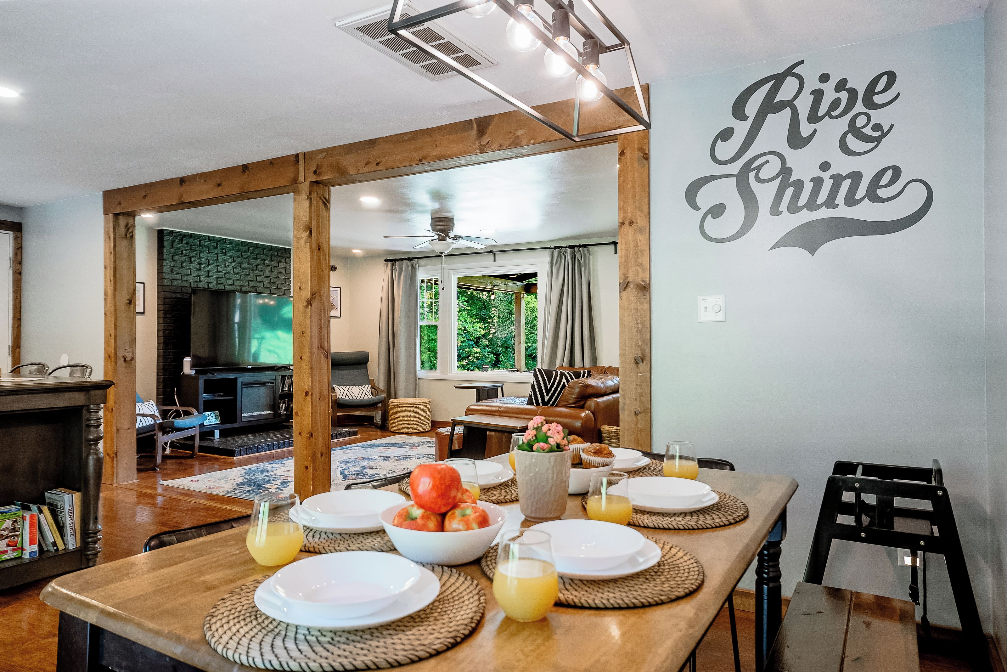 A dining area with a wooden table set for breakfast, a television in the background, and a wall with the words 'Rise & Shine'.