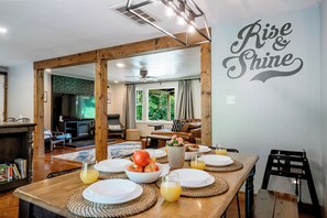 A dining area with a wooden table set for breakfast, a television in the background, and a wall with the words 'Rise & Shine'. - Family Fun Hocking Hills Retreat, 2 Decks, Game Rm (Logan)