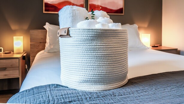 A neatly made bed with a large woven basket filled with white towels on a hotel room. - Family Fun Hocking Hills Retreat, 2 Decks, Game Rm (Logan)