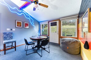 Una habitación con ventilador de techo, una mesa con sillas, un sillón de peluche y cuadros en la pared.