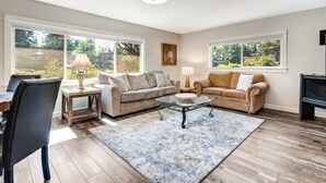 A living room with two sofas, a coffee table, a TV, and large windows. - Riverfront Cottage Just Steps From Deschutes River (Bend)