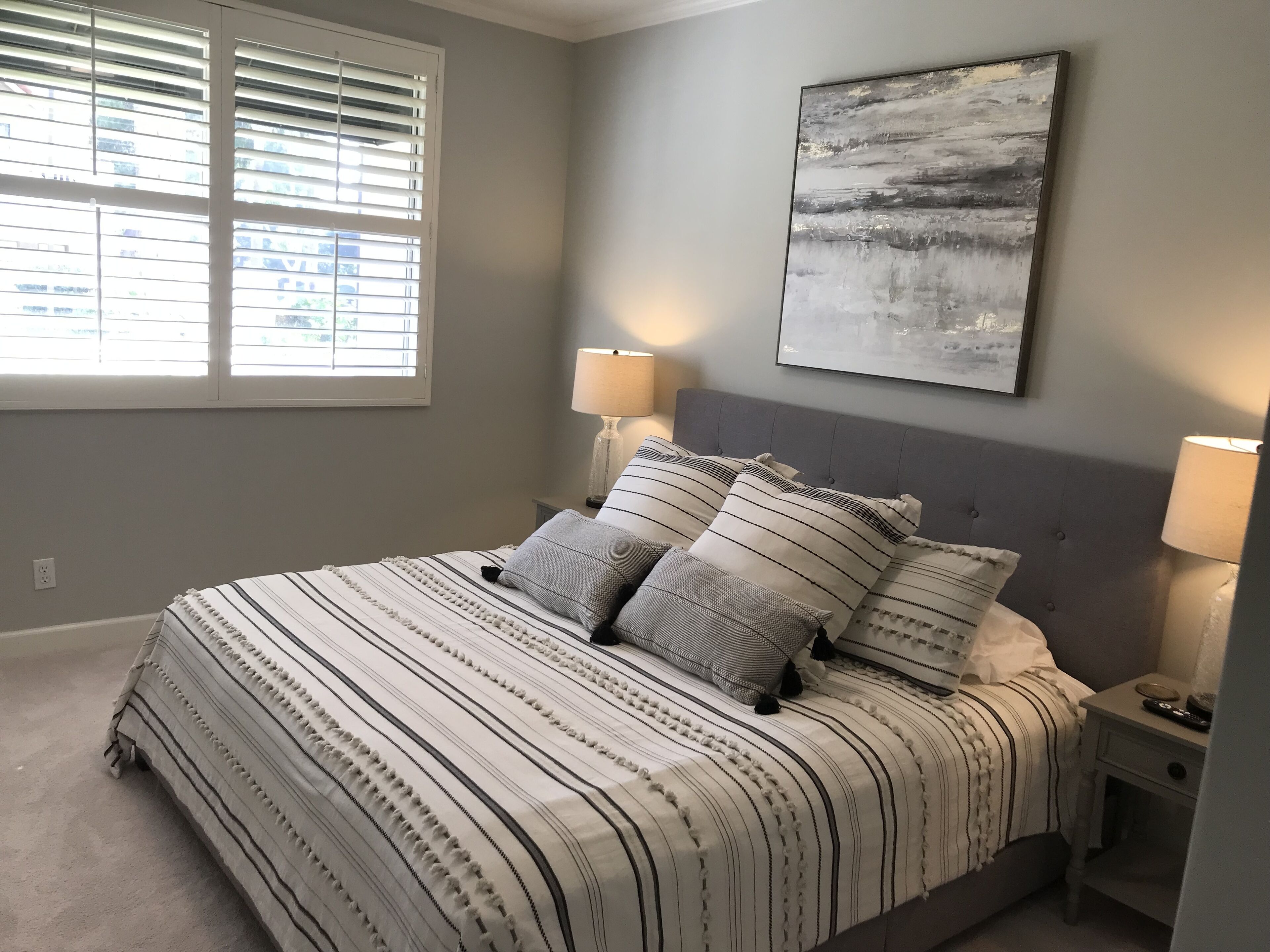 A neatly made bed with striped bedding and multiple pillows, a bedside table with a lamp, and a large abstract painting on the wall.