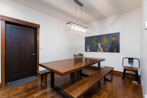 A dining area with a wooden table, benches, a hanging light fixture, a door, and a wall-mounted painting.