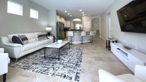 A modern living room with a sofa, coffee table, and TV. There is a kitchen area in the background with a bar and seating. - Crisp & Pristine Pool & Spa & Game Room Home! Solterra Resort! (Davenport)
