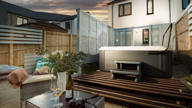 Une terrasse extérieure accueillante, dotée d’un jacuzzi, de meubles en osier et d’une table en verre sur laquelle sont posés des verres à vin et un appareil photo.