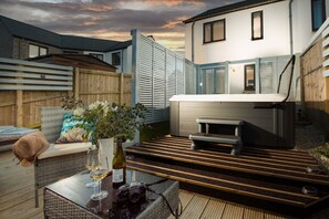 Un patio exterior acogedor con jacuzzi, mobiliario de mimbre y una mesa de vidrio con copas de vino y una cámara.