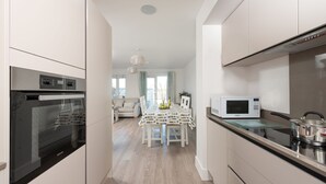 Une cuisine moderne dotée d’appareils électroménagers en acier inoxydable, un coin repas avec une table dressée pour deux personnes, et une vue sur le salon en arrière-plan.
