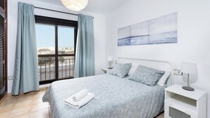 A bedroom with a bed, a nightstand, a lamp, a window with curtains, and a painting on the wall. - Home2Book Sea Side El Medano (El Medano)