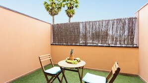 A small outdoor seating area with two wooden chairs and a round table, a pineapple, and a fruit bowl on a green artificial turf. - Home2Book Sea Side El Medano (El Medano)