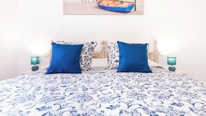 A bed with a blue and white patterned bedspread, two blue pillows, a matching pillow with a paisley design, and a framed picture of a boat on the wall. - Home2Book Jable Beach Apartment (Morro Jable)